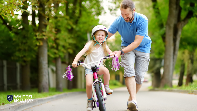 A father helps his young daughter ride a bike, smiling and guiding her along a tree-lined path.