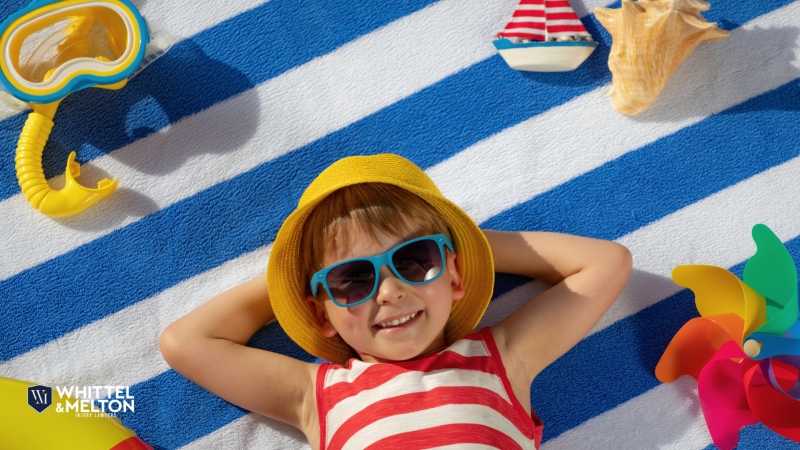 A young boy wearing sunglasses and a yellow hat smiles while lying on a blue and white striped towel surrounded by summer beach toys.